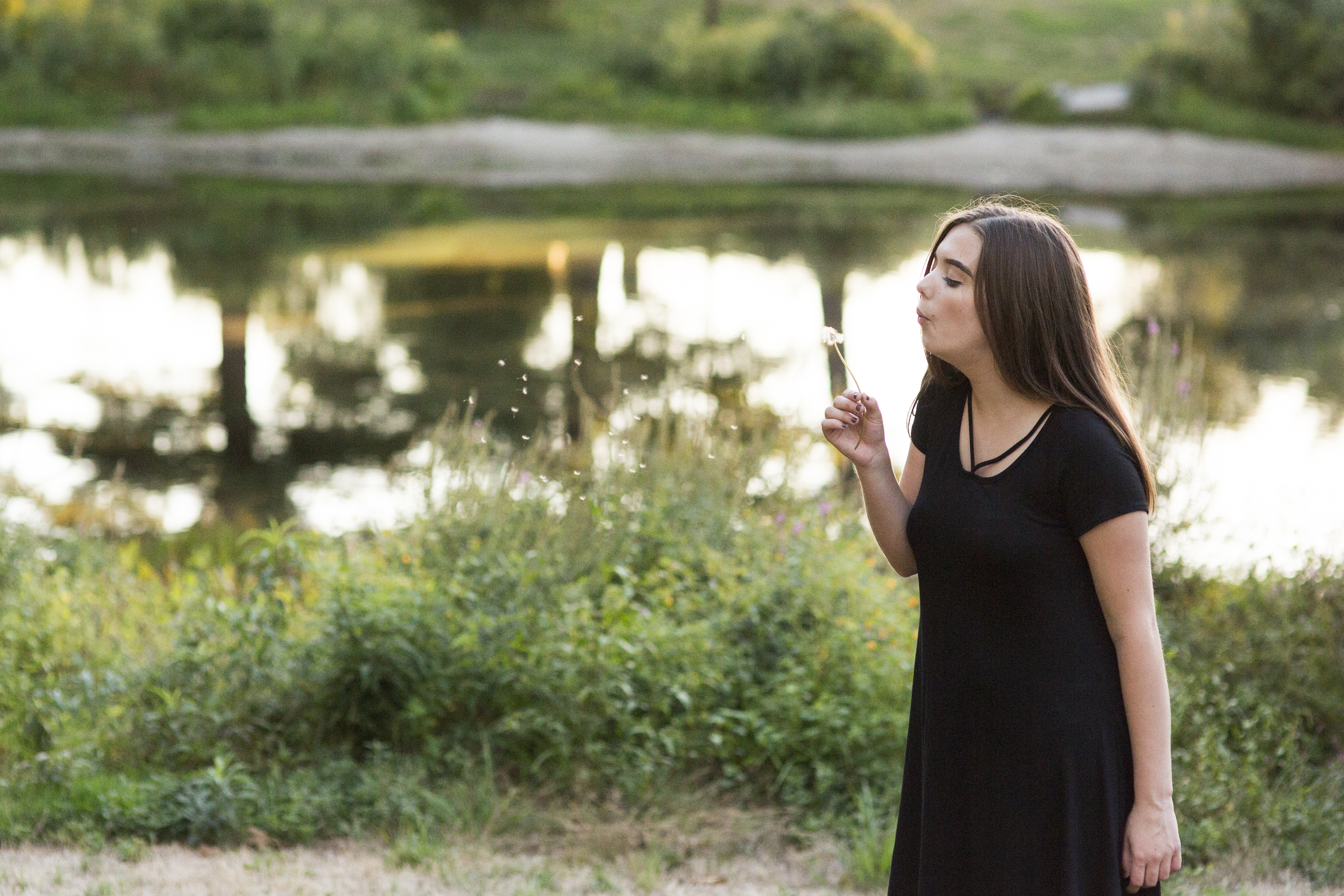 girl blows dandelion in front of lake golden hour senior photo