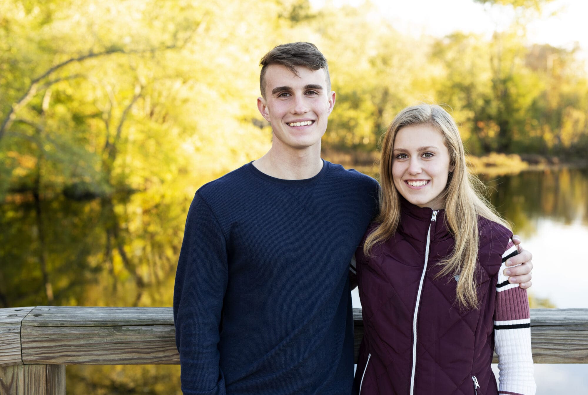 twin senior boy and girl siblings standing on a wooden bridge over the water with fall leaves